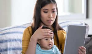 A mom uses her palm to check her infant's temperature as she uses a tablet computer to meet with a doctor for a virtual visit