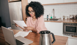 A woman sits at her kitchen table with a pot of coffee and a laptop as she tries to make sense of a doctor's bill