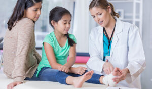 A doctor in a lab coat examines a young girl's foot during an office visit while the child's mom observes