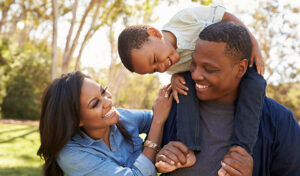 As a family visits a park, a young son sits on his dad's shoulders, while his mom embraces her husband and smiles.