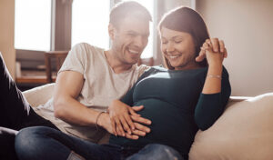 A young man places his hand on his wife's belly to feel a kicking baby as the couple relaxes on a sofa.