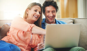 A couple sits together on a sofa at home, completing a health risk assessment on a laptop computer.